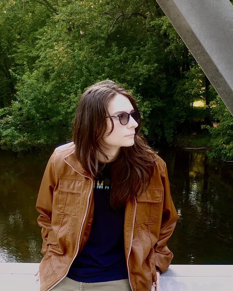 Violet Love in a tan leather jacket and sunglasses, leaning on the rail of a bridge over a wooded river.